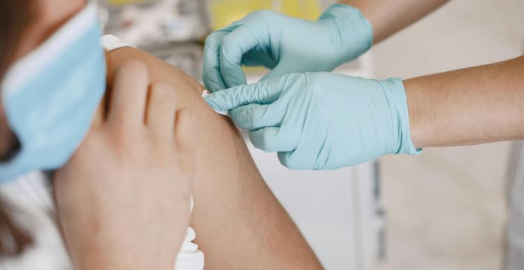 Close-up of a healthcare worker wearing light blue medical gloves administering an injection into a person’s upper arm while the person wears a face mask, in a clinical indoor setting.