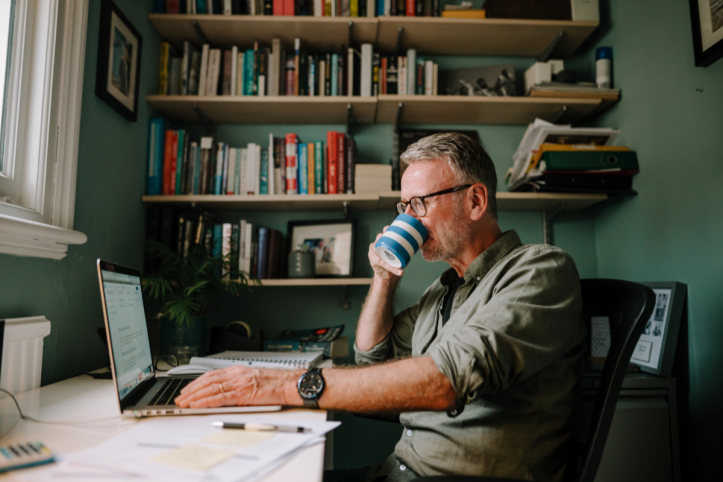 a man sitting in his home office filling out a survey on his laptop while drinking a cup of tea or coffee