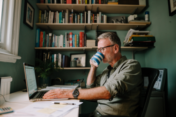 a man sitting in his home office filling out a survey on his laptop while drinking a cup of tea or coffee