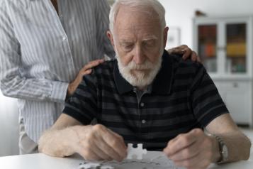 Older man with a beard completing a jigsaw with someone behind, helping him with their hands on his shoulders.