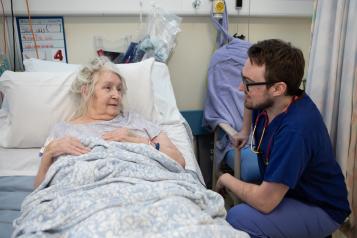 Older patient lying in a hospital bed speaking with a healthcare worker in scrubs and a stethoscope.