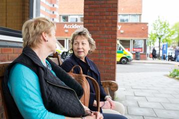 Two older women sat on a  bench talking outside a hospital