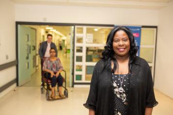 Woman in hospital smiling at the camera with a man pushing a woman in a wheelchair in the background