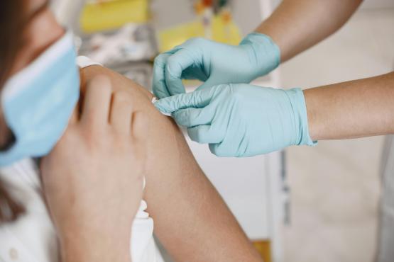 Close-up of a healthcare worker wearing light blue medical gloves administering an injection into a person’s upper arm while the person wears a face mask, in a clinical indoor setting.