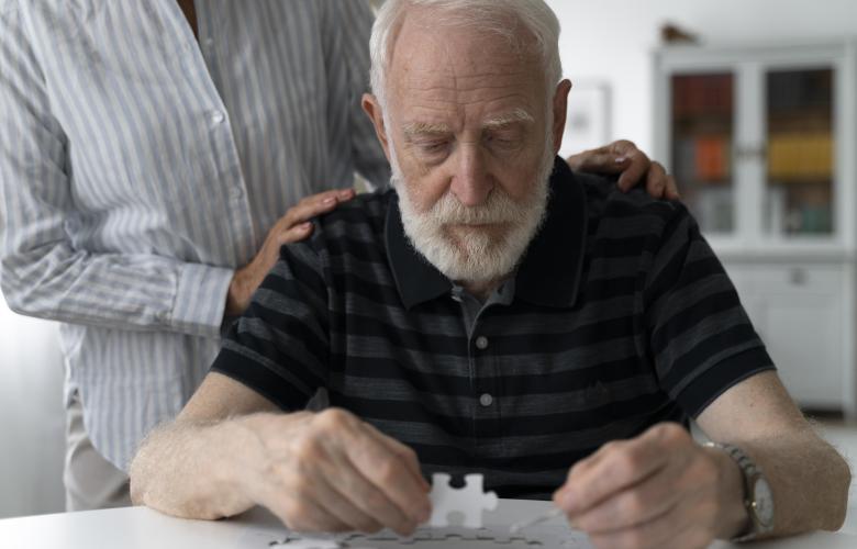 Older man with a beard completing a jigsaw with someone behind, helping him with their hands on his shoulders.