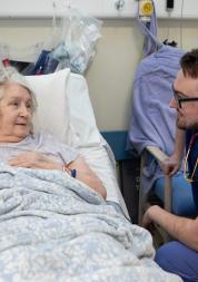 Older patient lying in a hospital bed speaking with a healthcare worker in scrubs and a stethoscope.
